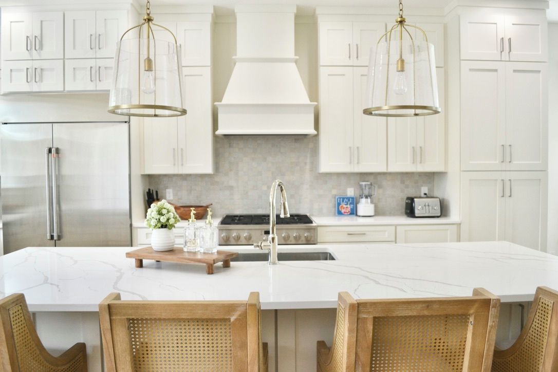 A kitchen with white cabinets, a marble island, wicker chairs, and pendant lights.