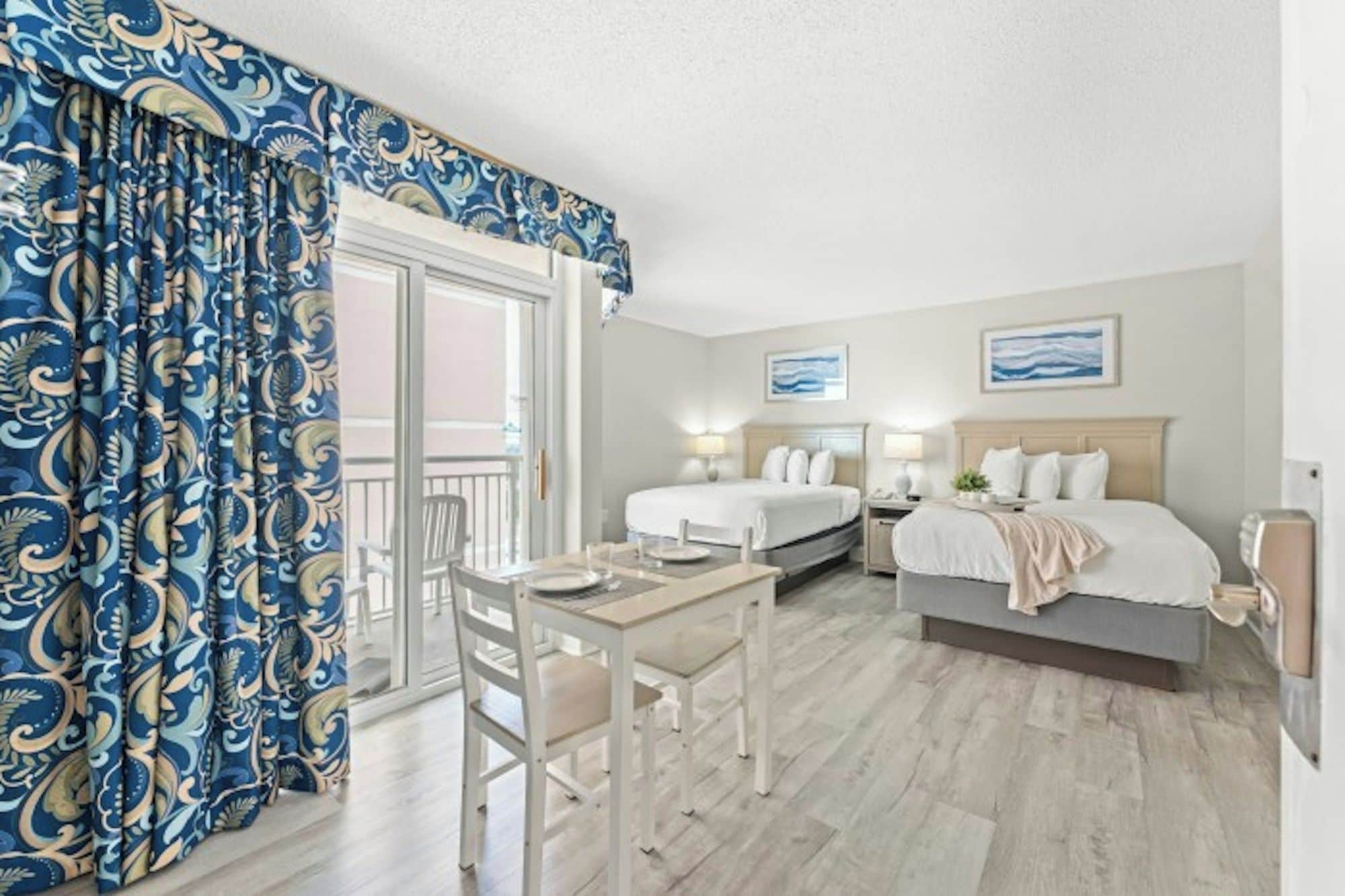 A hotel room with a balcony, two beds, a dining table, and a chair.