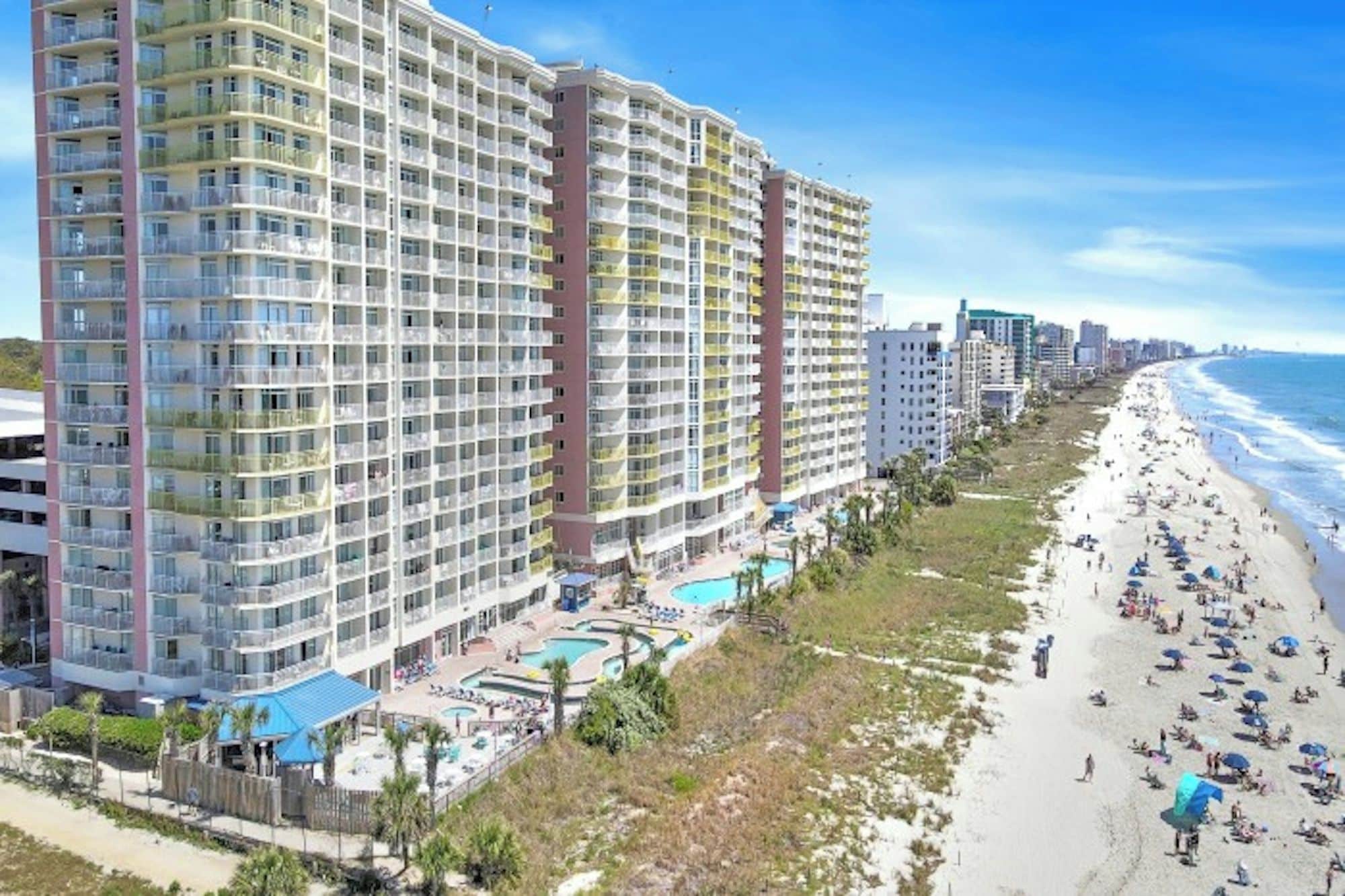 A beachfront hotel with multiple floors, a pool area, and a beach with umbrellas.