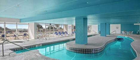 Una piscina interior con borde de azulejos, sillones de descanso y una zona cubierta con vistas al exterior.