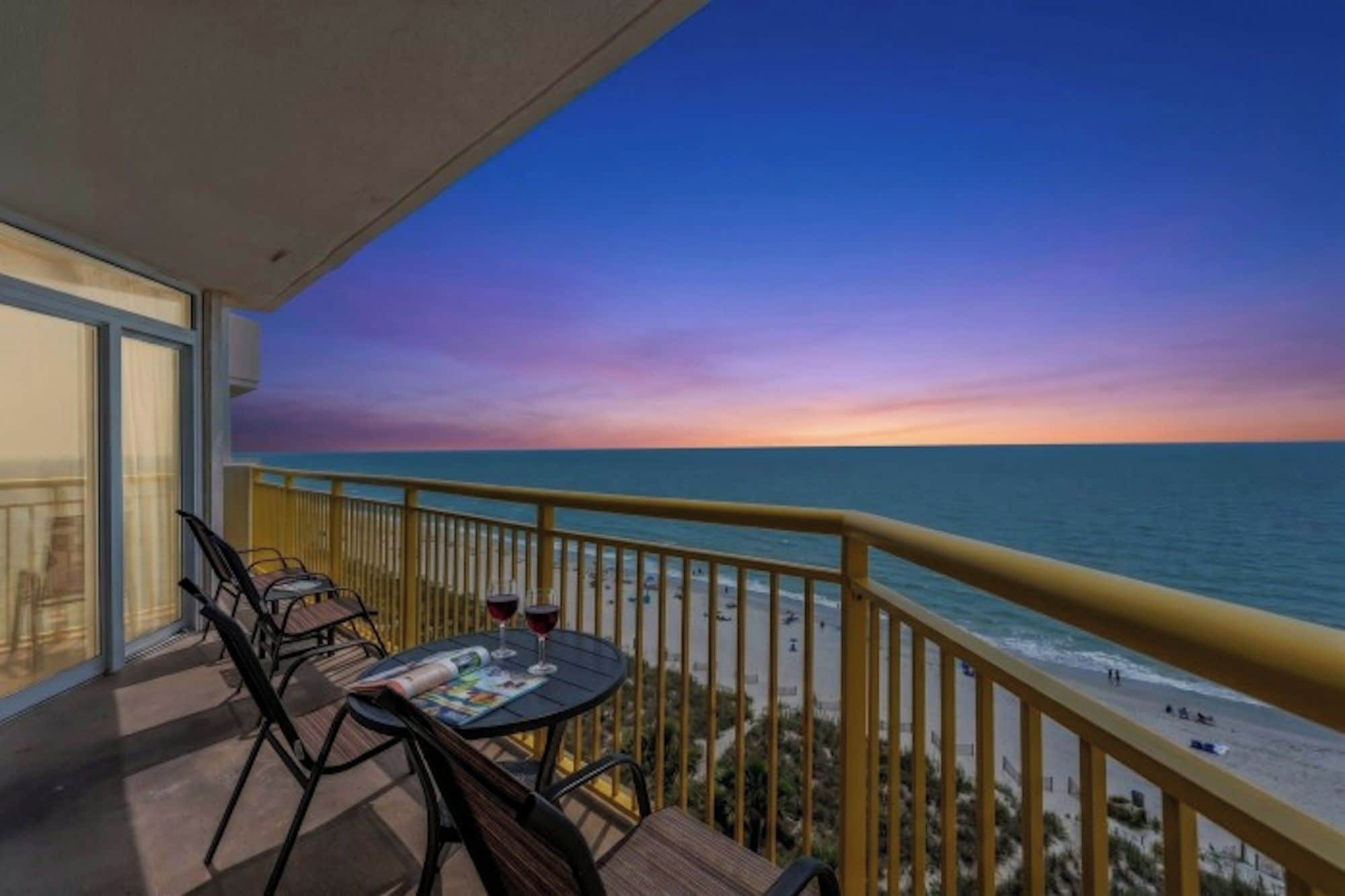 Een balkon met een tafel en stoelen, met uitzicht op een strand bij zonsondergang.