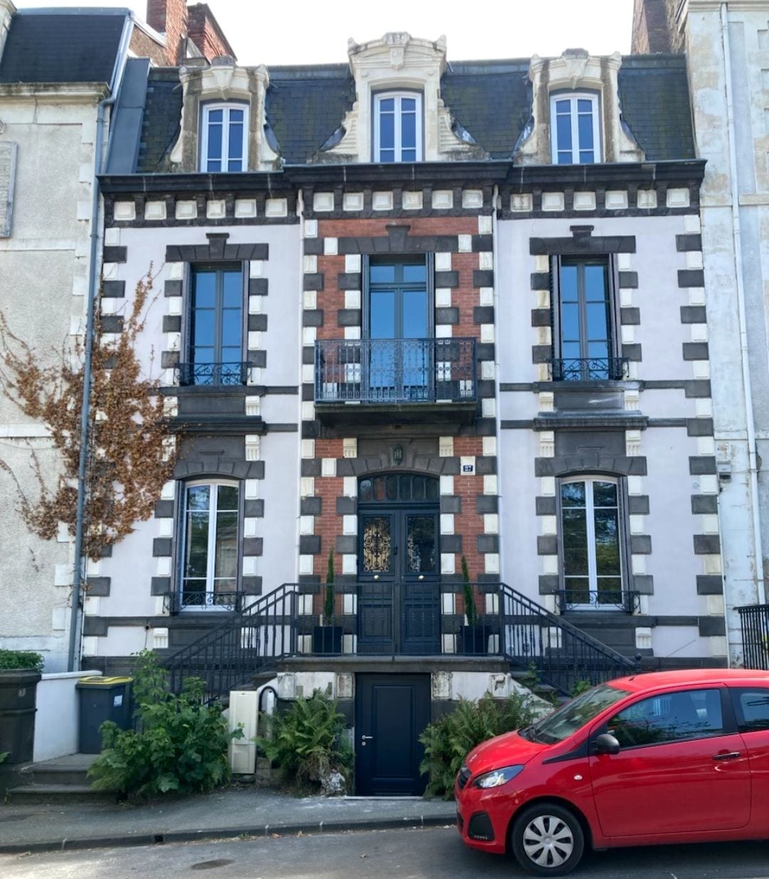Un edificio de varios pisos con una puerta negra, un balcón y un coche rojo estacionado adelante.
