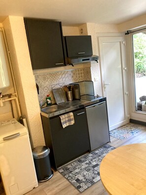 A compact kitchen with a built-in oven, a small refrigerator, and a coffee machine. There is a window with a view of greenery outside. - Cosy studio 27, à 400m des thermes (Chamalières)