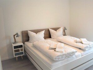 Ein ordentlich bezogenes Bett mit weißen Leinen, zwei Nachttischlampen und ein kleiner Nachttisch mit Schubladen.