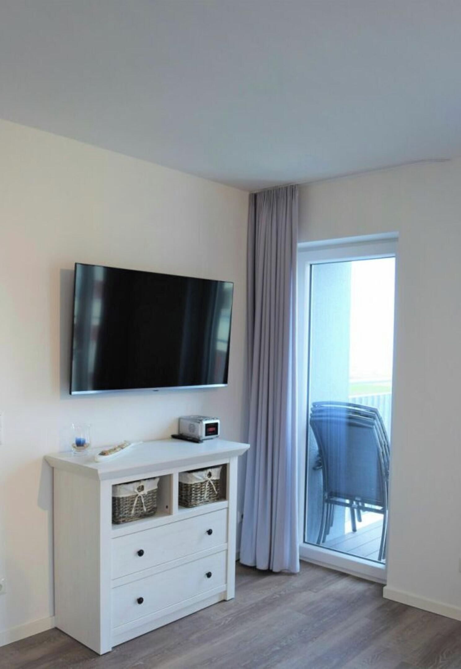 A modern room with a flat-screen TV mounted on the wall, a white dresser with drawers, and a sliding glass door leading to an outdoor area.