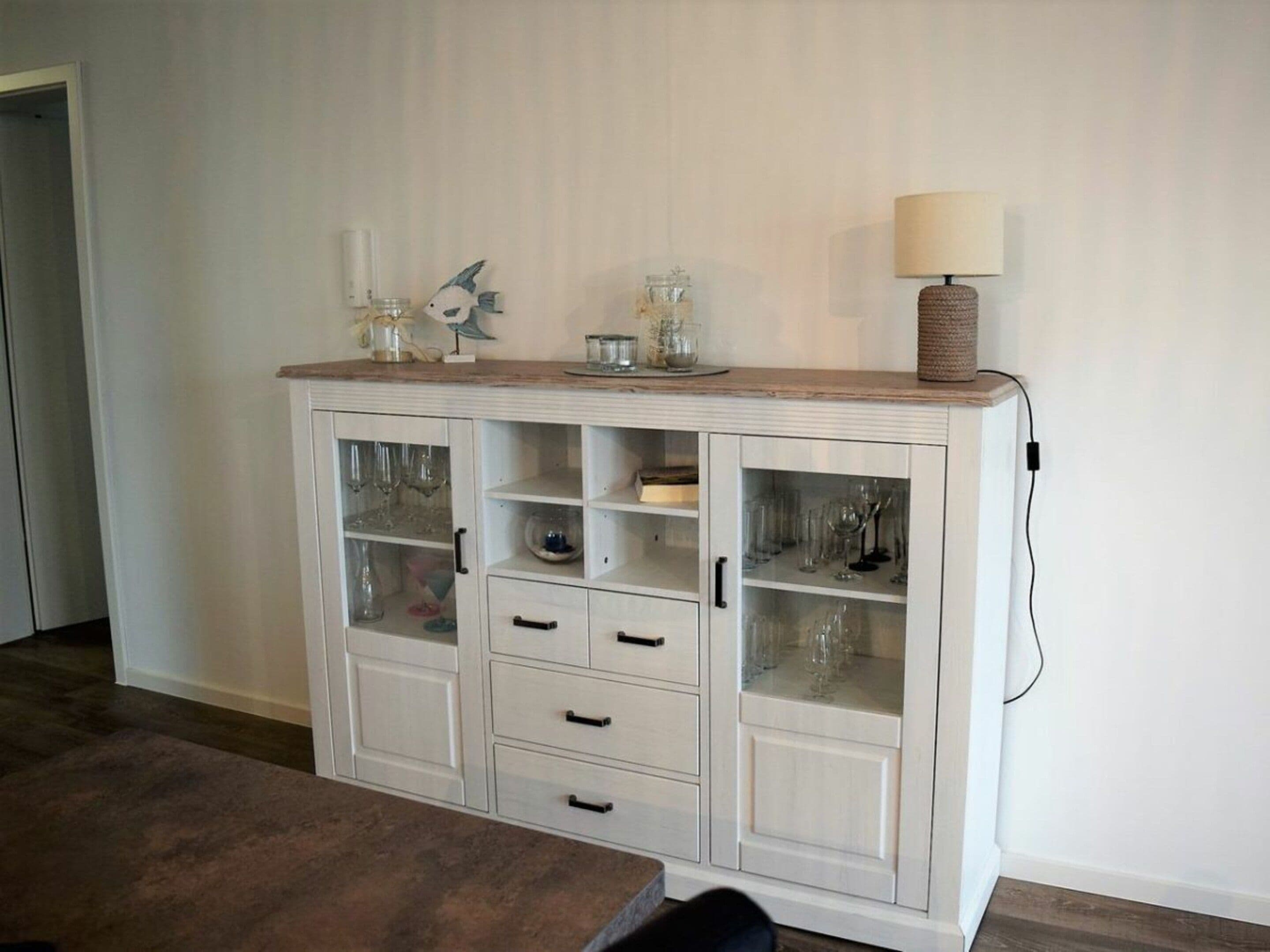 A white wooden cabinet with glass doors, drawers, and shelves, some containing glassware.