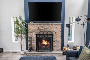 Ein Wohnzimmer mit Kamin aus Ziegelsteinen, einem Flachbildfernseher darüber, einer Zimmerpflanze und einem blauen Sofa.