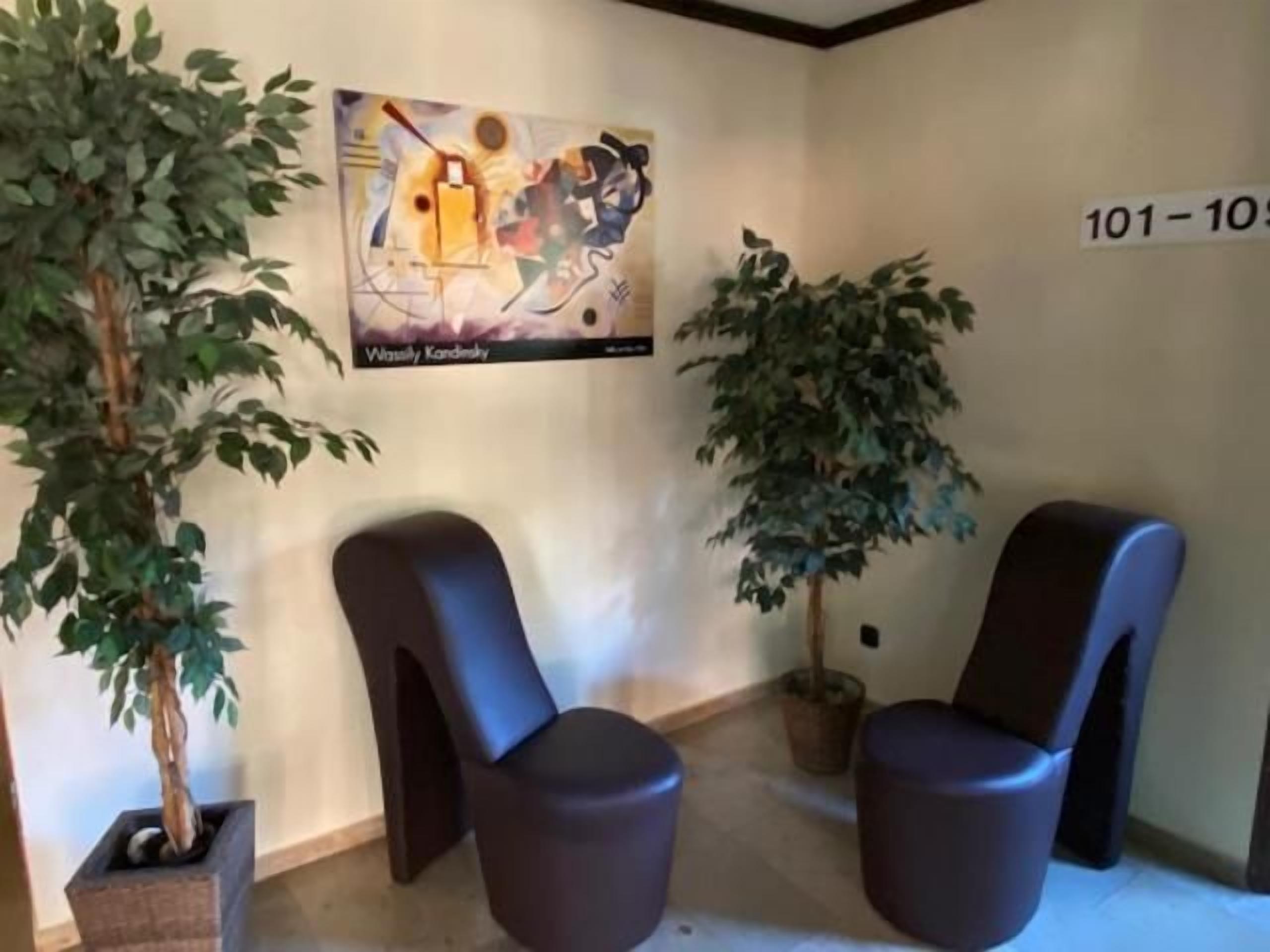 Ein Wartebereich mit zwei modernen Stühlen, einer Topfpflanze und einem an der Wand befestigten Kunstwerk.