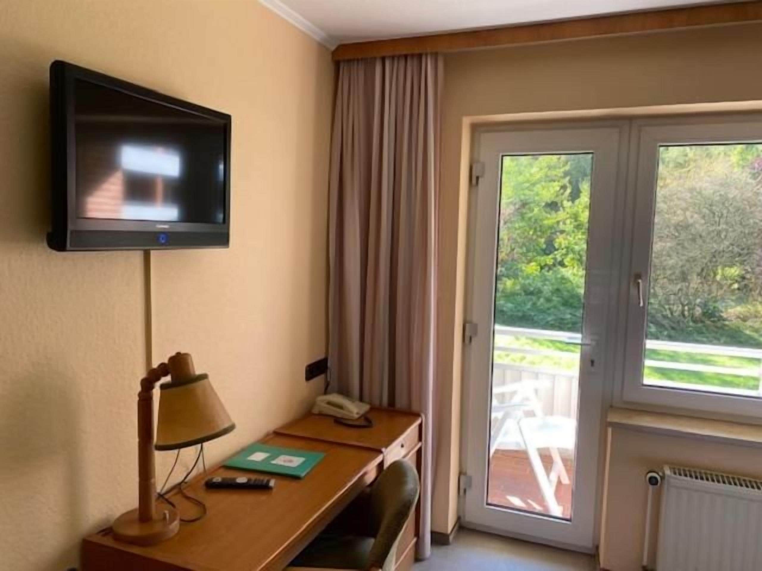 Ein Hotelzimmer mit einem an der Wand befestigten Fernseher, einem Schreibtisch mit Lampe, einem Telefon und Blick ins Freie durch eine Schiebetür.