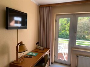 A hotel room with a television mounted on the wall, a desk with a lamp, a telephone, and a view of the outdoors through a sliding glass door. - Hotel Tanneneck (Bad Bramstedt)