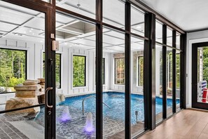 A modern indoor pool with clear glass walls, a stone feature, and a view of greenery outside. - Smoky Mountain Game Luxe Lodge (Gatlinburg)