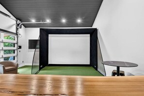 Une pièce avec une moquette verte, une table en bois, un écran noir en forme de filet et un mur blanc sur lequel est fixé un téléviseur.