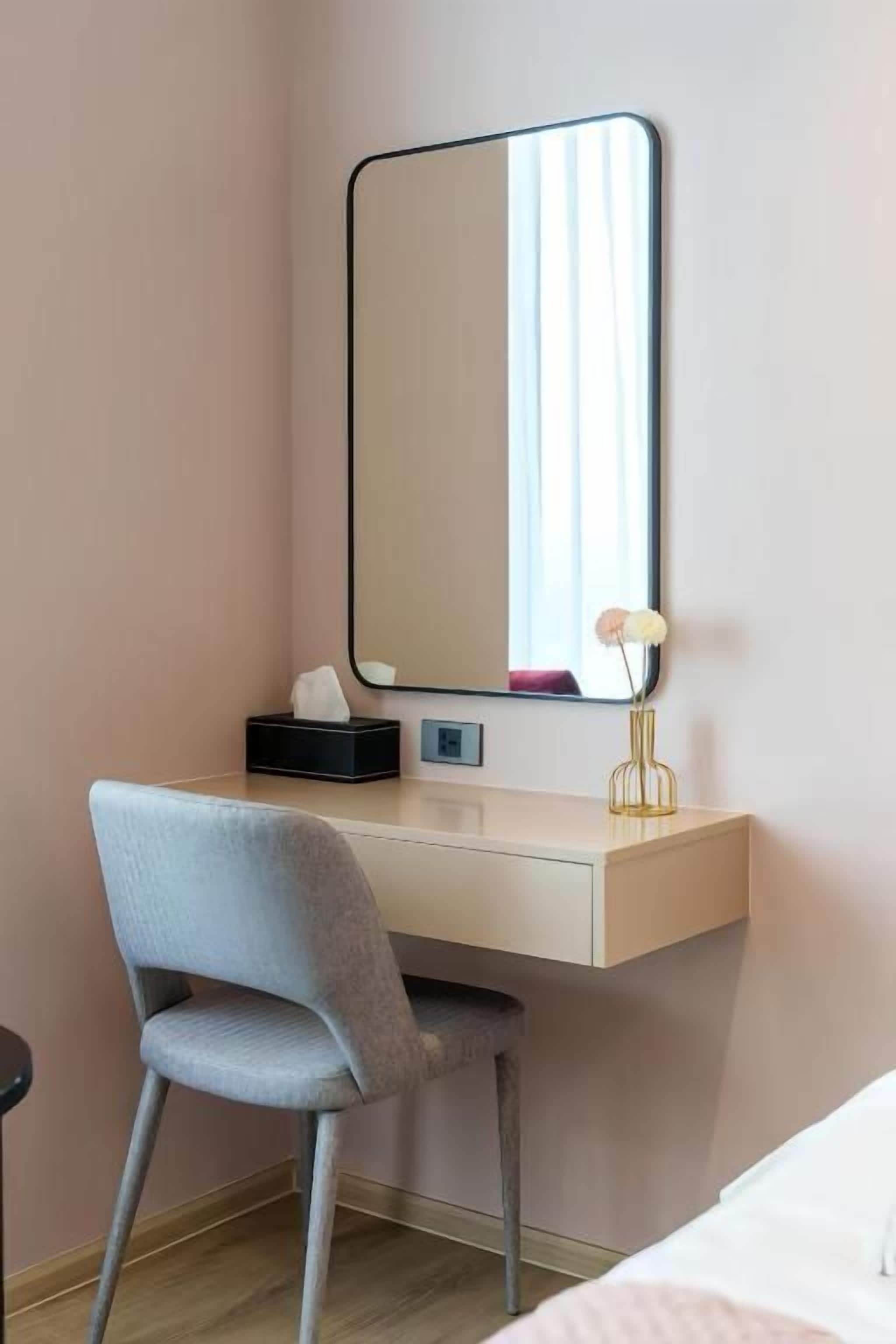 A hotel room with a desk, a chair, a mirror, and a bedside table.