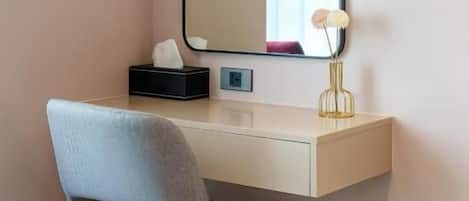 A hotel room with a desk, a chair, a mirror, and a bedside table.