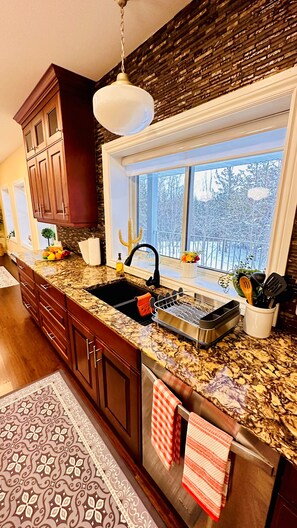 A kitchen with a granite countertop, stainless steel sink, wooden cabinets, a hanging light fixture, and a window with a view of trees. - Cozy Retreat On The Eastside Estate (sleep14) (Greater Napanee)