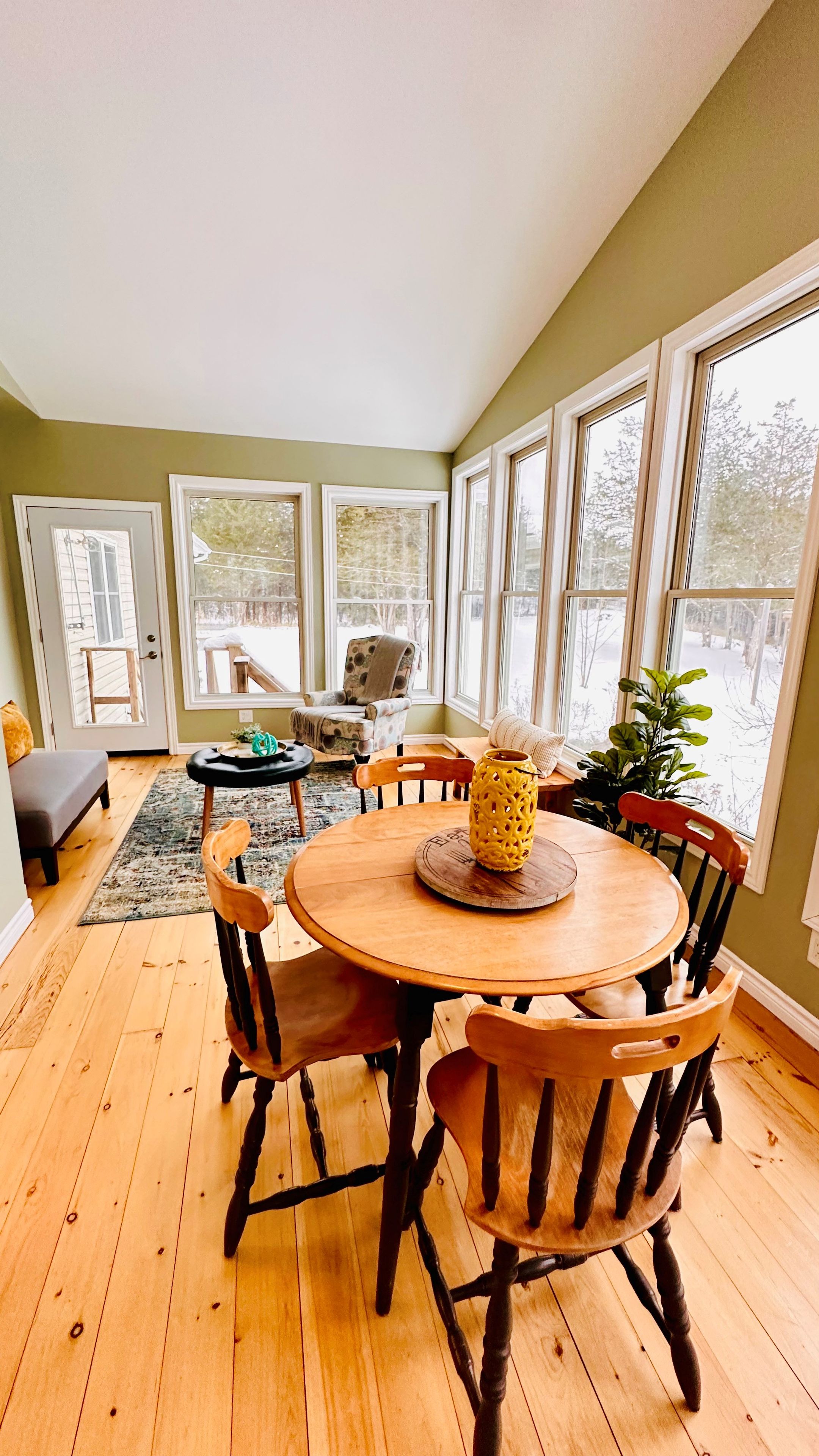 Un comedor con una mesa redonda de madera y sillas, un jarrón decorativo amarillo y vistas a una terraza nevada a través de grandes ventanales.