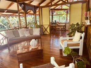 A spacious room with a wooden floor, a sofa, a dining table, and chairs. There are potted plants and a view of greenery outside. - Casa Caribeña (Limón)