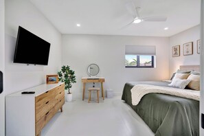 A bedroom with a bed, a dresser, a desk with a mirror, a television mounted on the wall, and a ceiling fan. - Tiki on the Green - Pool, Bar, and Putting Green (Palm Beach Gardens)