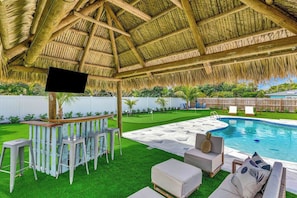 An outdoor pool area with a thatched roof, bar with stools, and lounge seating. - Tiki on the Green - Pool, Bar, and Putting Green (Palm Beach Gardens)