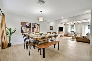 A modern dining area with a wooden table and chairs, a bench, and a hanging light fixture. - Tiki on the Green - Pool, Bar, and Putting Green (Palm Beach Gardens)