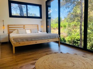 Une chambre à coucher avec un lit en bois, une table de chevet, une lampe fixée au mur et une grande fenêtre donnant sur des arbres et de la verdure.
