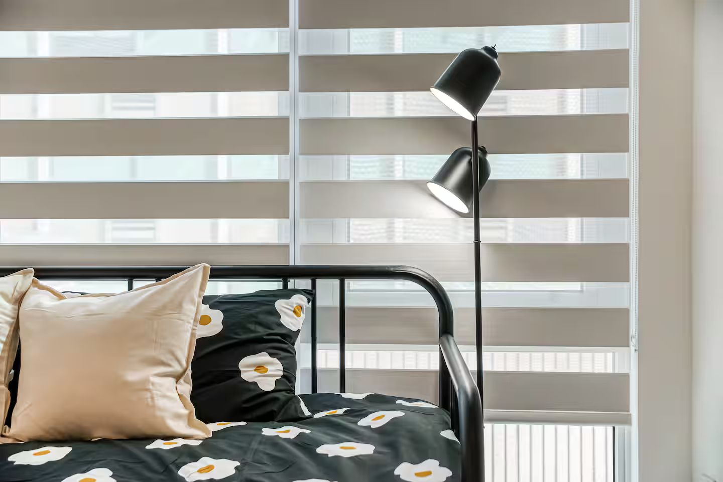 A bed with a black metal frame, a pillow, and a blanket with a white and black floral pattern. There is a black floor lamp beside the bed.