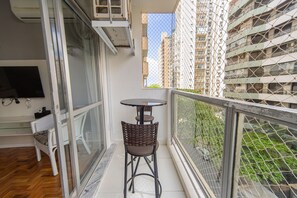 A balcony with a table and chair, a TV, and a view of tall buildings. - Apt for 5 w/ AC 150m from Pitangueiras Beach (Guarujá)