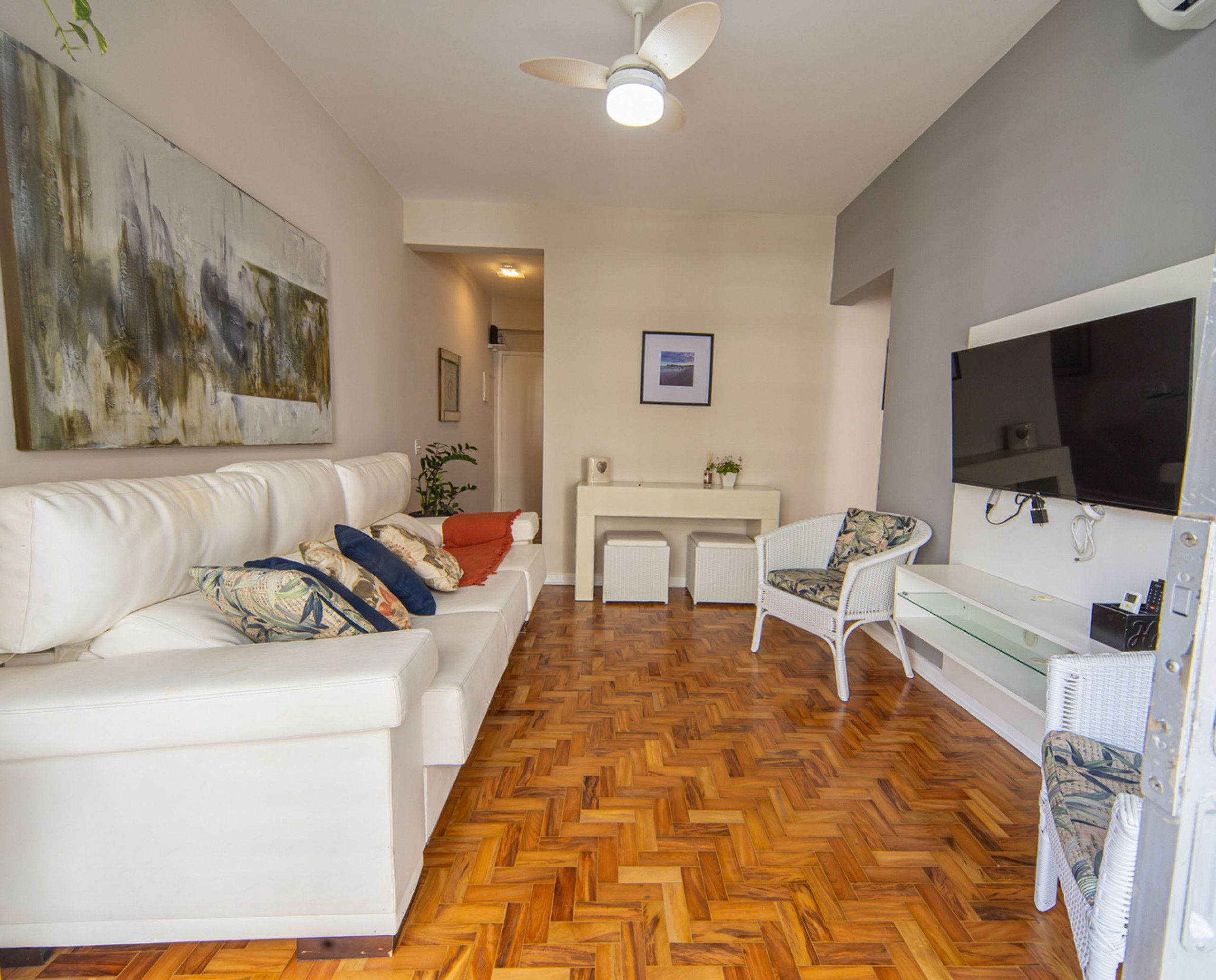 Ein Wohnzimmer mit weißem Sofa, Holzboden im Fischgrätmuster, Deckenventilator, einem an der Wand montierten Fernseher und einem Gemälde an der Wand.