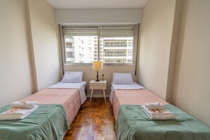 Quarto de hotel pequeno com duas camas de solteiro, mesa de cabeceira com abajur e janela com persianas.