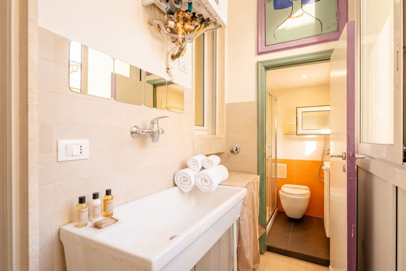 Un bagno con un lavandino bianco, due specchi, un portasciugamani con asciugamani e un WC visibile attraverso una porta aperta.