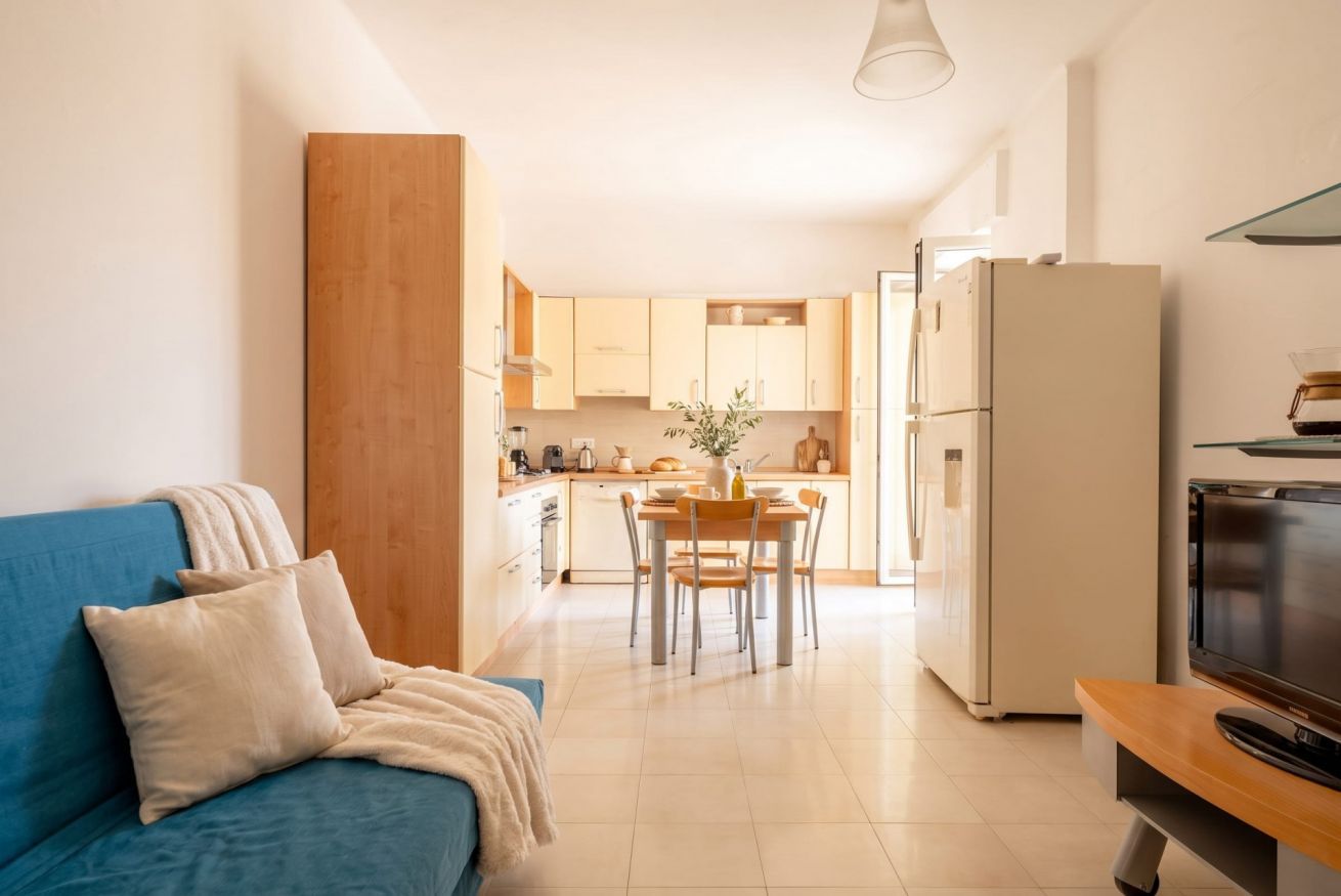 Un soggiorno moderno con divano, zona pranzo con tavolo e sedie, cucina con mobili ed elettrodomestici, e una televisione su un mobile.
