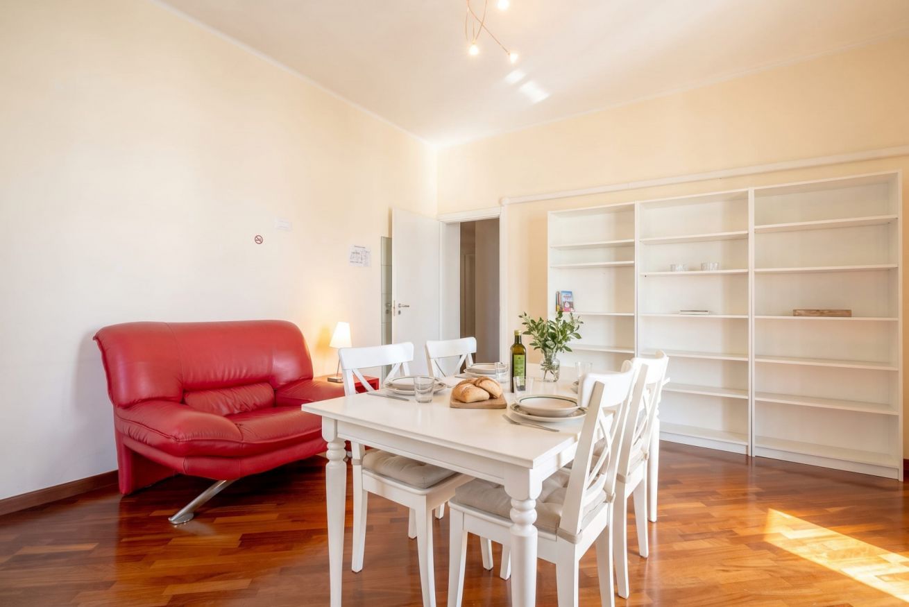 Un'area pranzo con un tavolo e sedie bianche, un divano rosso e un mobile contenitore bianco addossato alla parete.