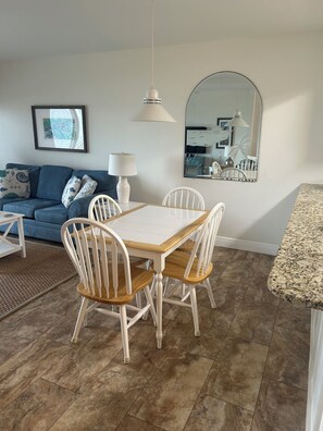 A dining area with a wooden table and white chairs, a blue sofa with decorative pillows, a white lamp, and a large mirror on the wall. - Beautiful 1 bedroom condominium steps away from the sugar white beach! (Fort Walton Beach)