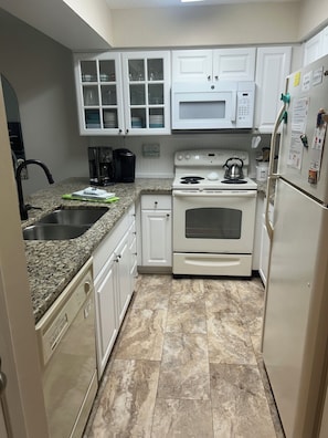 A compact kitchen with white cabinetry, a stainless steel sink, a microwave, an oven, and a refrigerator. - Beautiful 1 bedroom condominium steps away from the sugar white beach! (Fort Walton Beach)