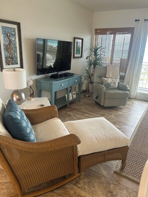 A living room with a television, a blue entertainment unit, wicker furniture, and a large window with blinds. - Beautiful 1 bedroom condominium steps away from the sugar white beach! (Fort Walton Beach)