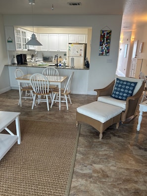 Een moderne keuken met witte kasten, een eethoek met een houten tafel en stoelen, en een zithoek met een rieten bank en een gekleurd kussen.