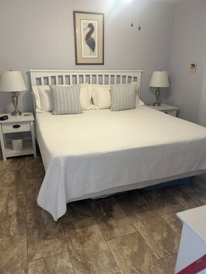 A neatly made bed with a quilted white cover, two bedside tables with lamps, a framed picture on the wall, and a tiled floor. - Beautiful 1 bedroom condominium steps away from the sugar white beach! (Fort Walton Beach)