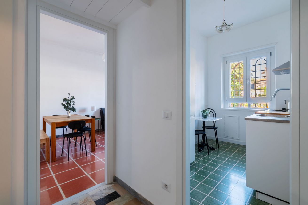 A hallway leading to a dining area with a table and chairs, a kitchen with a window, and a refrigerator.