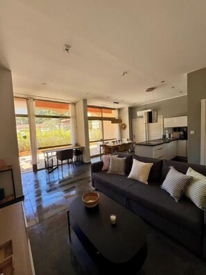A modern living room with a dark sofa, a coffee table, and a view of the kitchen and dining area. - Apartment, big terrace, pool, walk to sea (Sanary-sur-Mer)