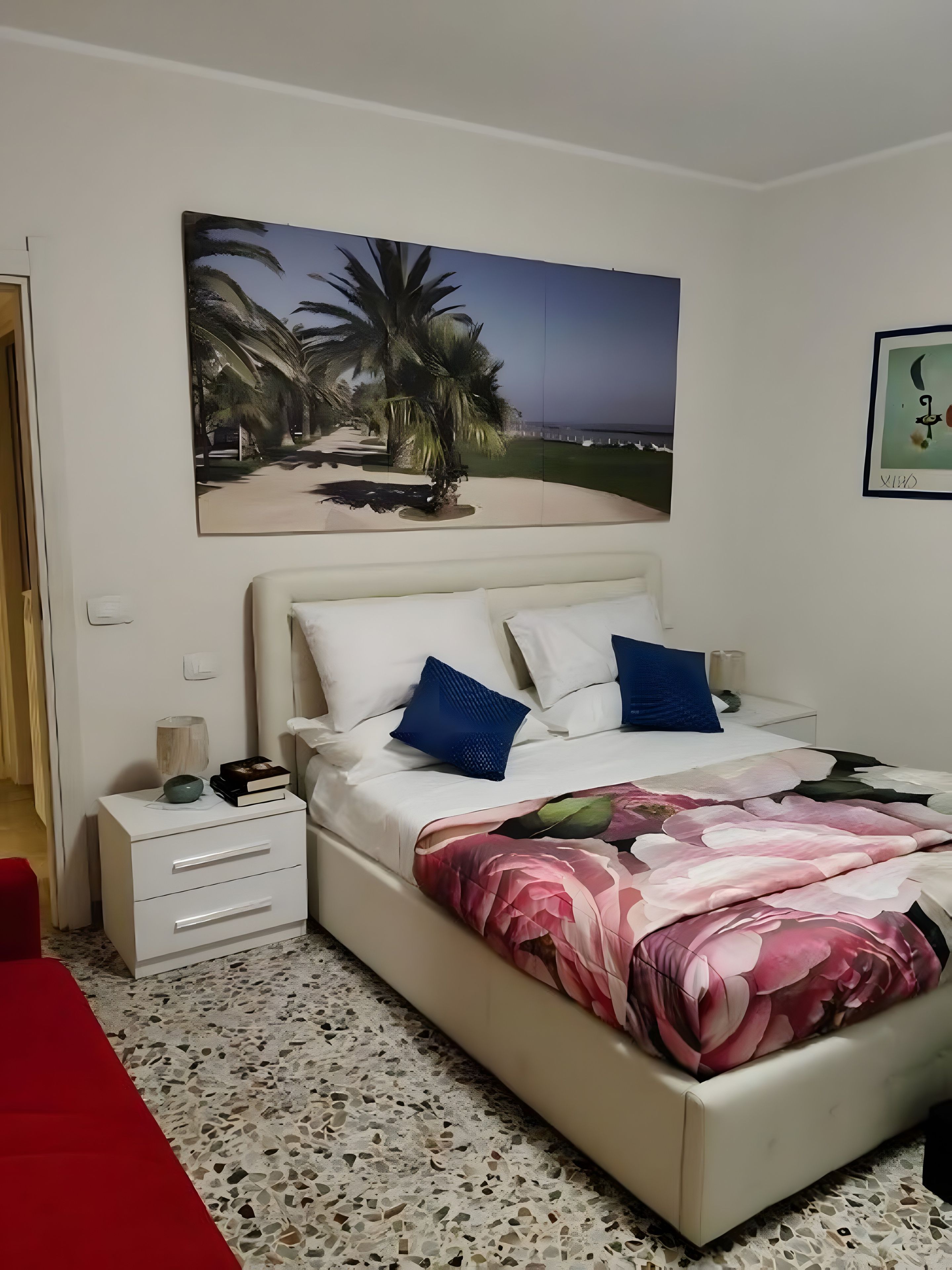 Un dormitorio con cama, mesita de noche, un cuadro y vista a palmeras.