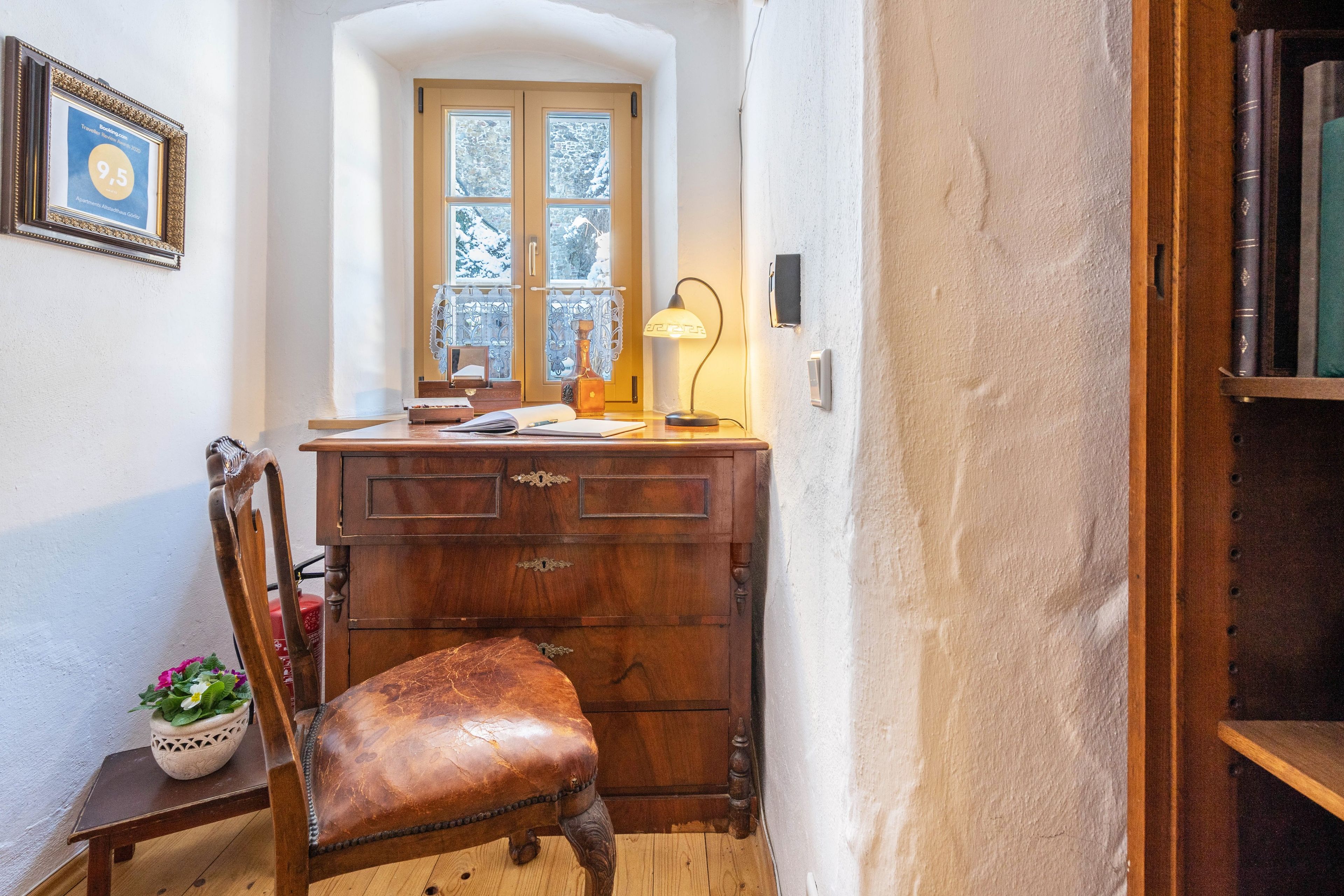 Quarto de canto com escrivaninha de madeira, uma cadeira, janela com vista para árvores cobertas de neve e um certificado emoldurado na parede.