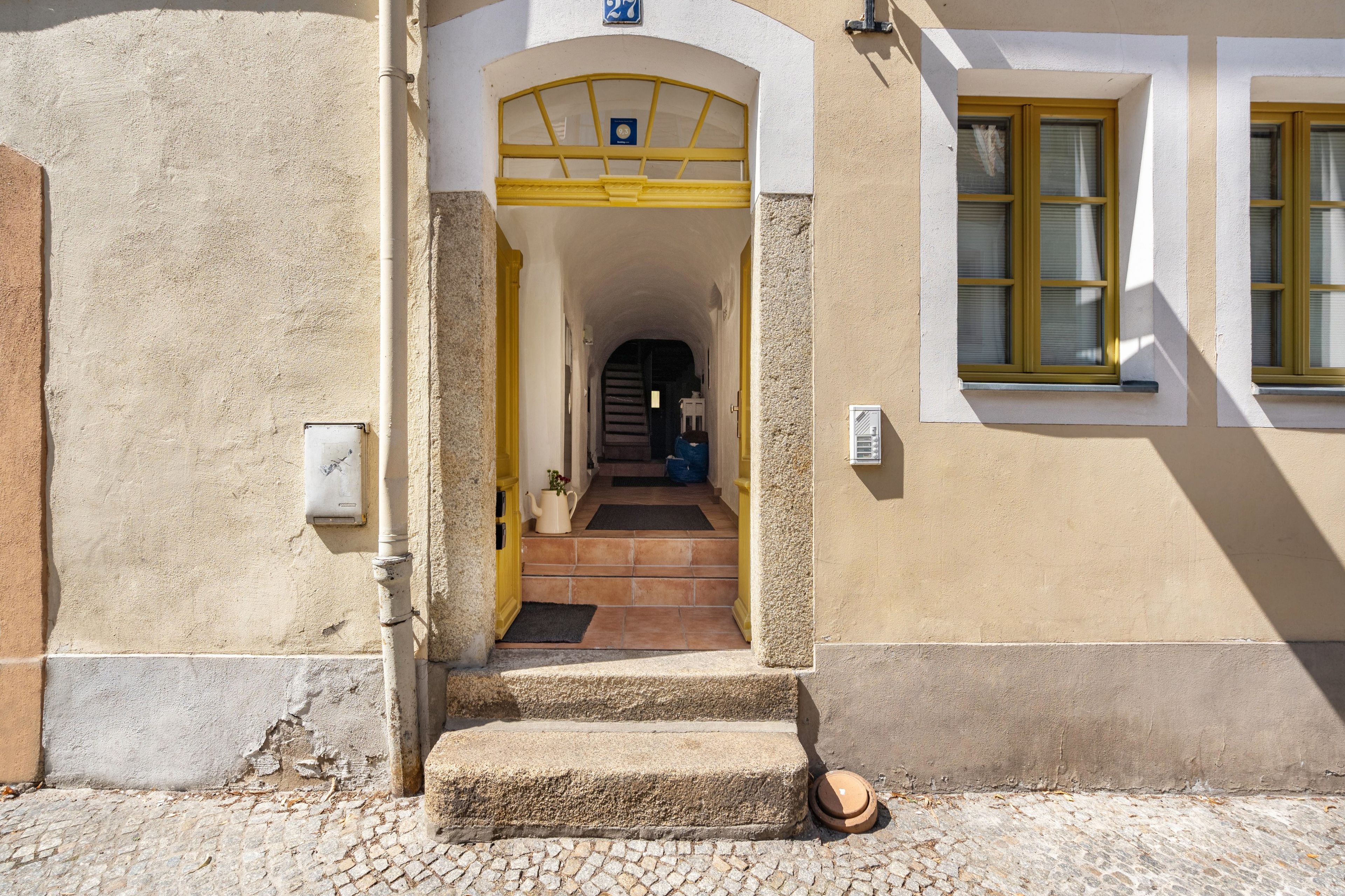 A building entrance with a yellow door, a tiled pathway leading to the entrance, and a window with yellow shutters.