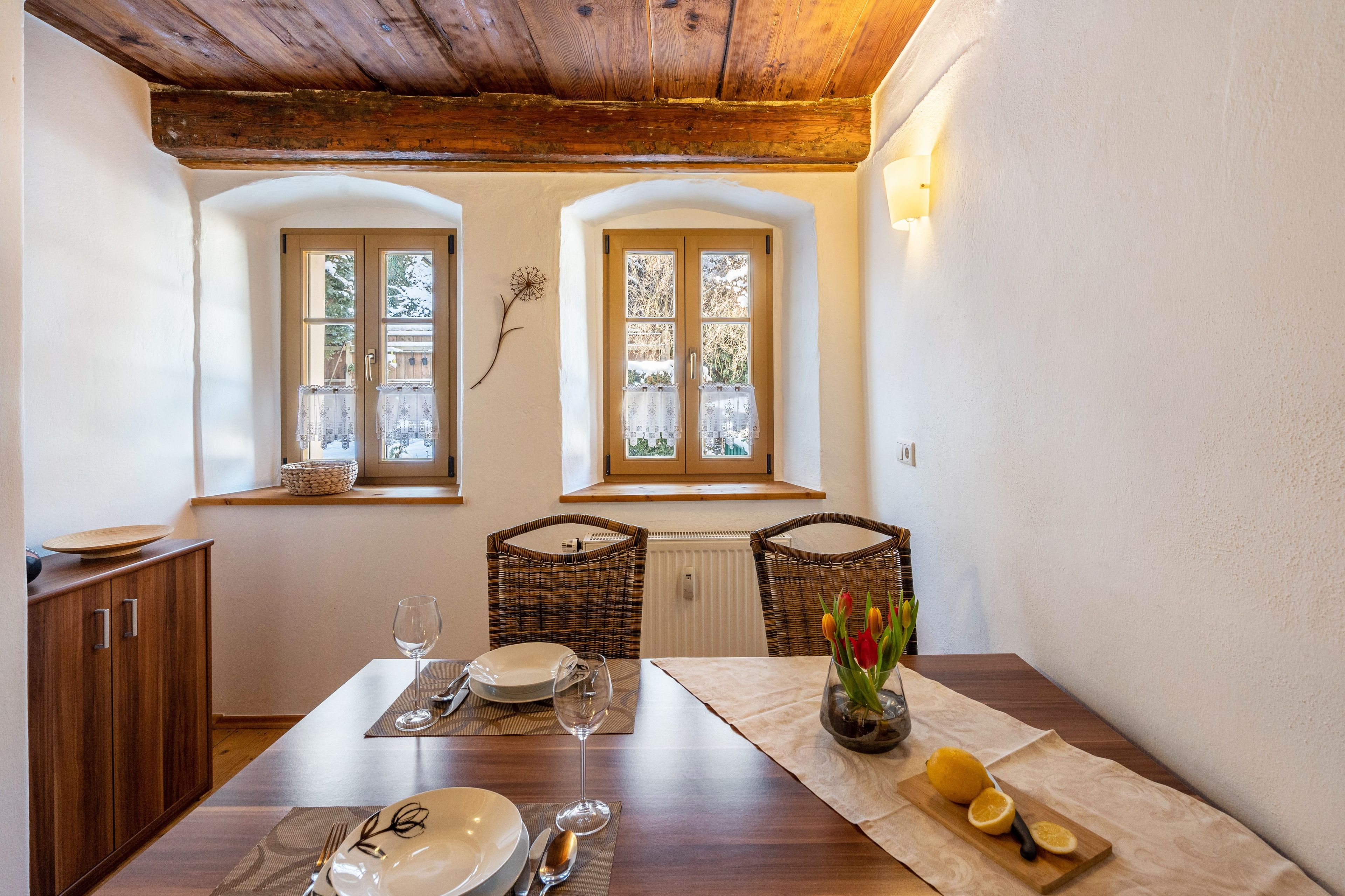 A dining area with a wooden table set for two, two wicker chairs, a vase with flowers, and a window with a view of trees.
