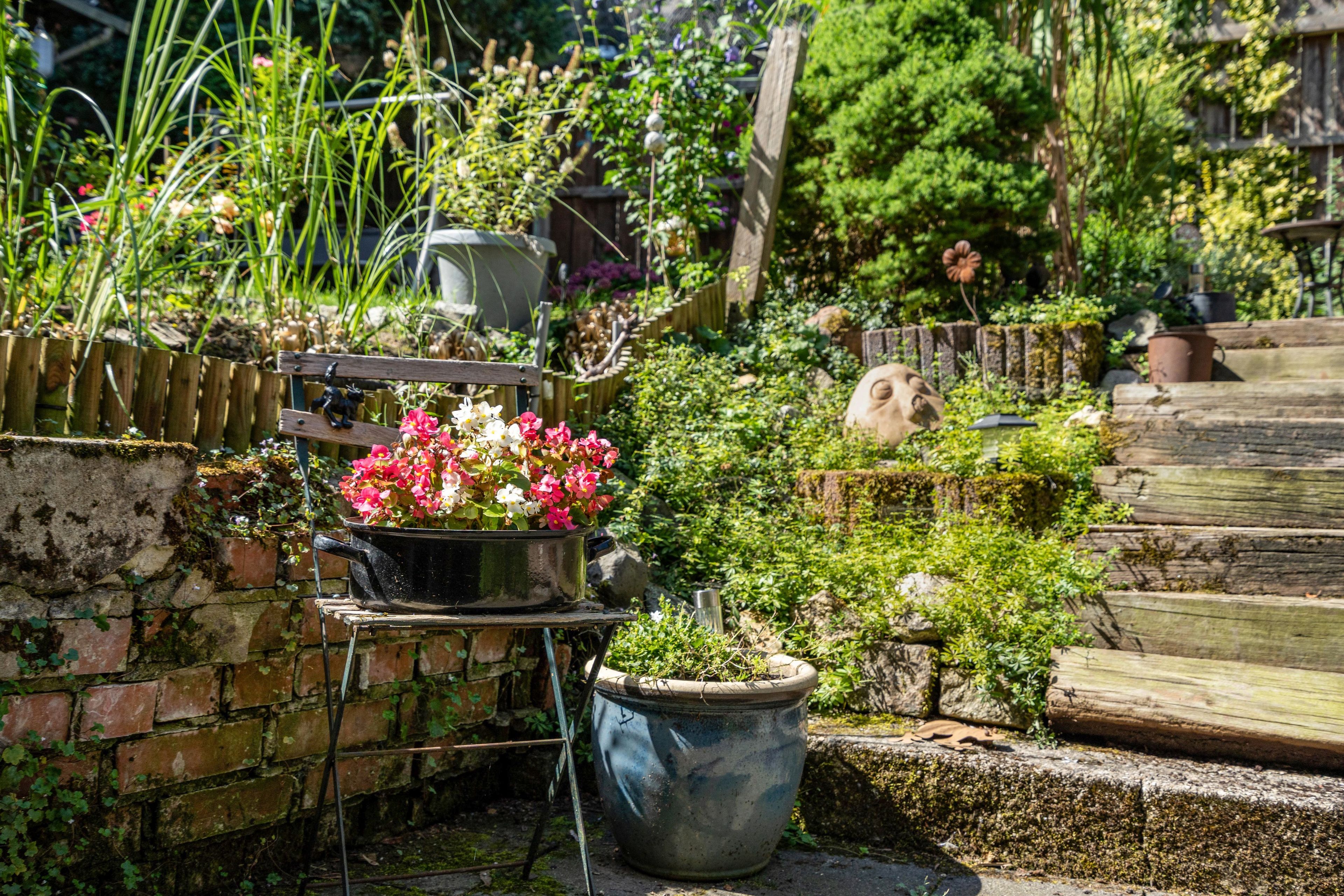 A garden with a variety of plants and flowers, a metal pot on a stand, and a wooden fence.