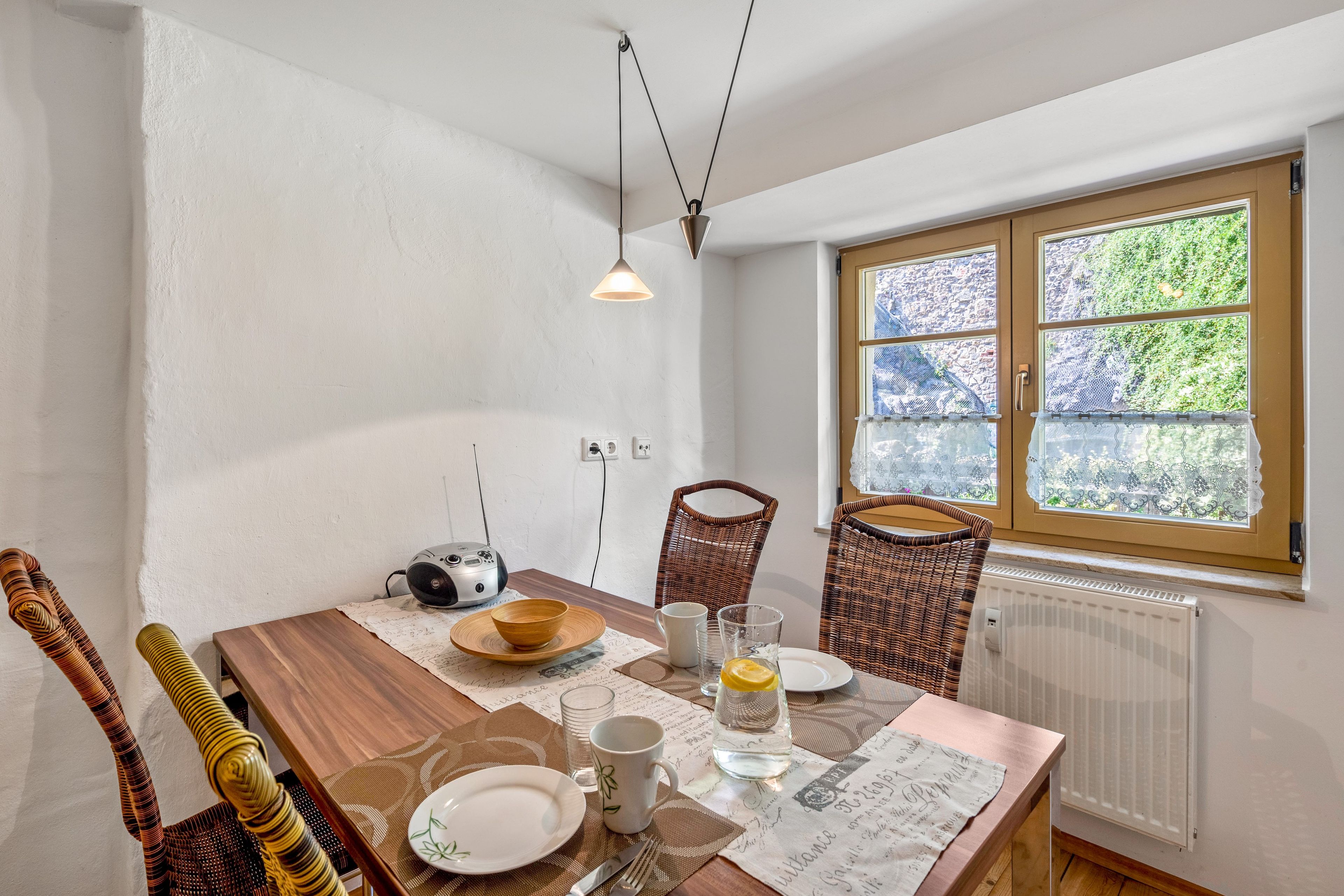 A dining area with a wooden table, chairs, a radio, and a window with a view of greenery.