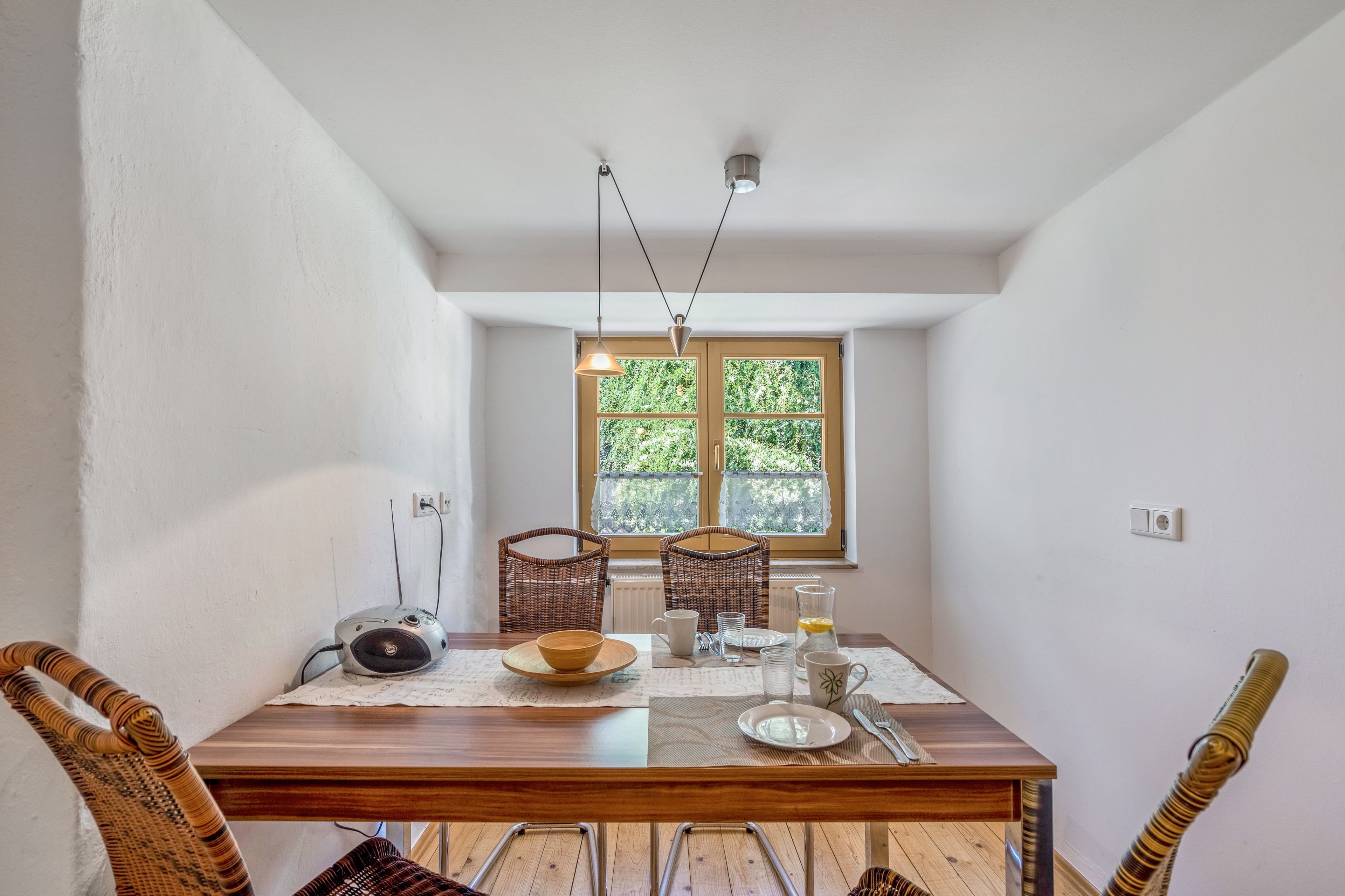 A dining area with a wooden table, wicker chairs, a window with a view of greenery, and pendant lighting.
