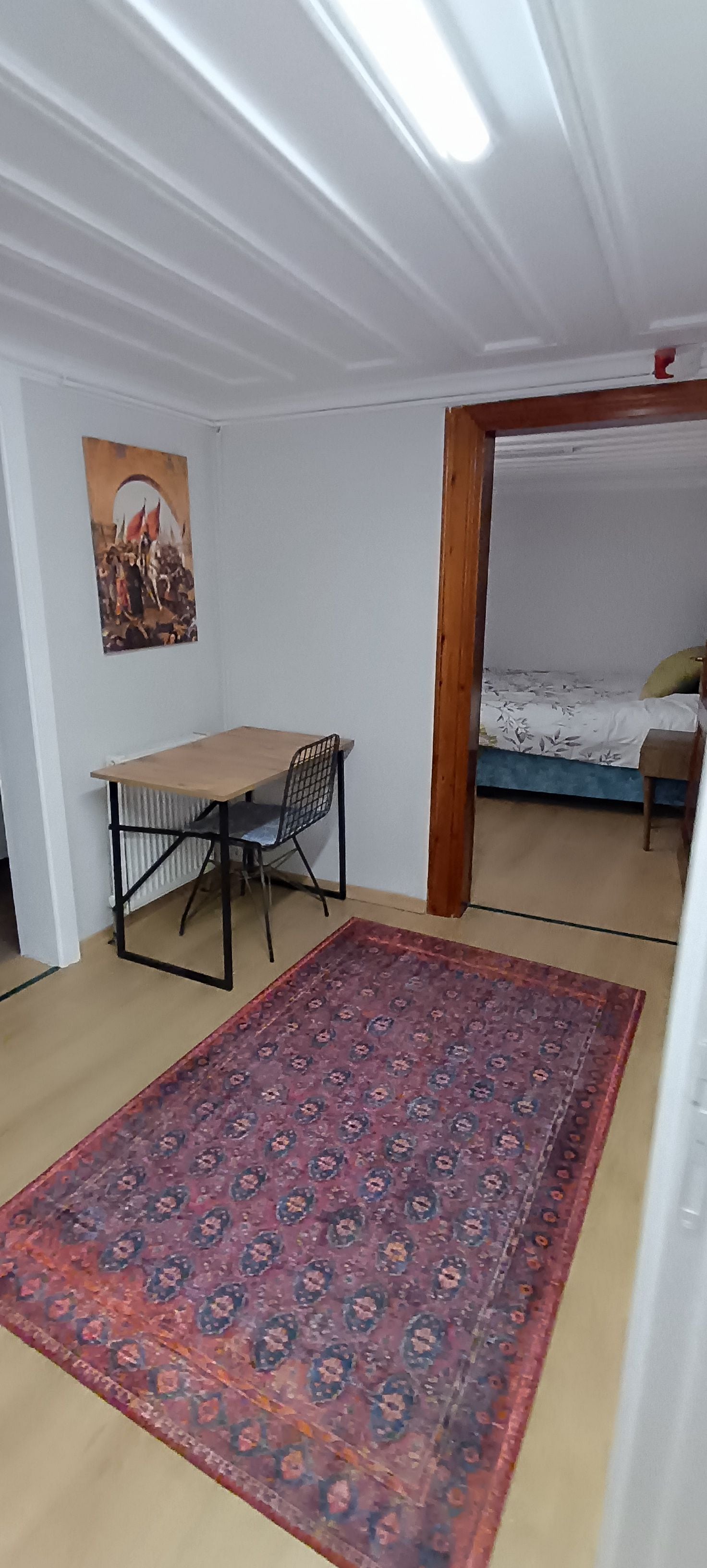 A room with a bed, a desk with a chair, a patterned rug, and a painting on the wall.