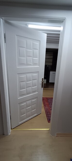 Uma porta branca com painel texturizado, dando acesso a um quarto com cozinha visível e um tapete vermelho no chão.