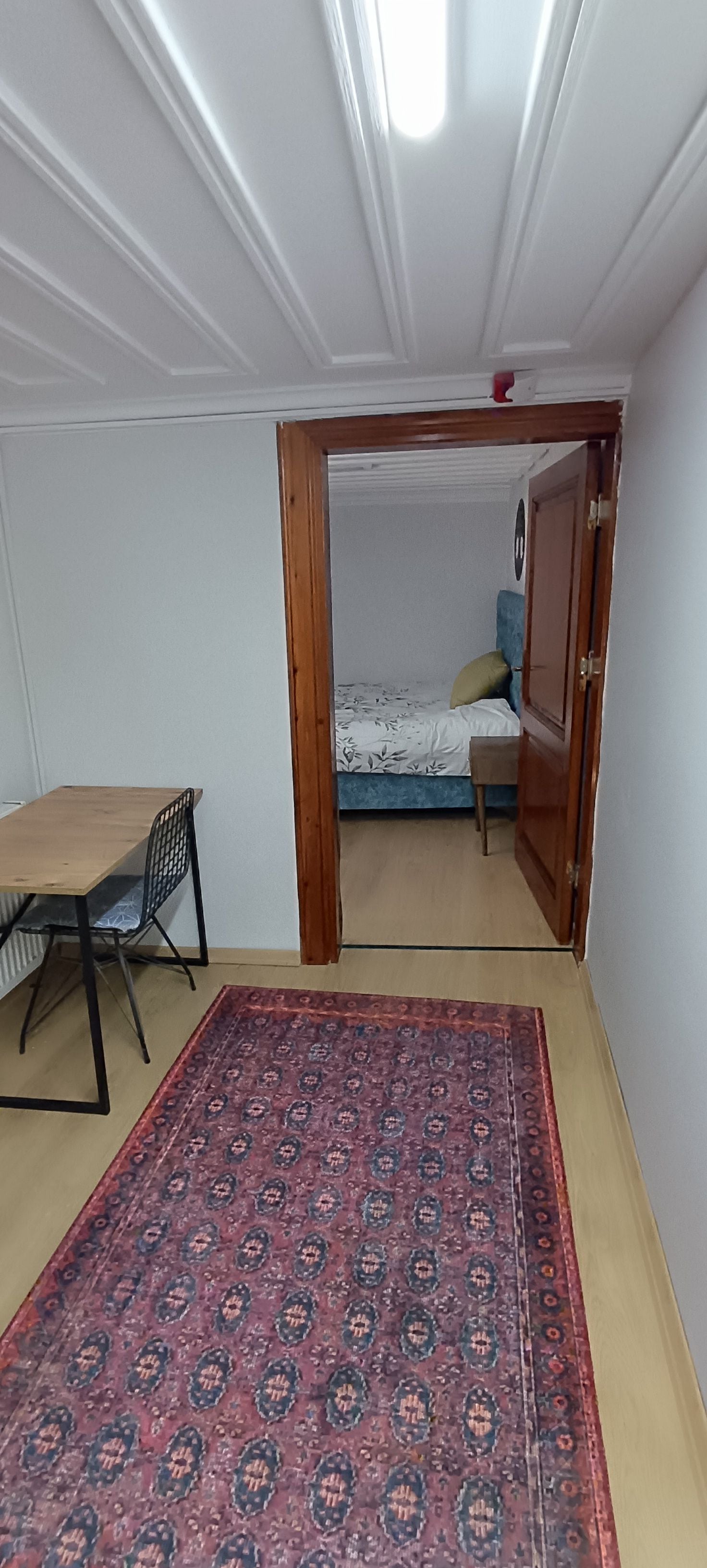Quarto com mesa de madeira, tapete estampado, cama com cabeceira azul e porta para outro cômodo.