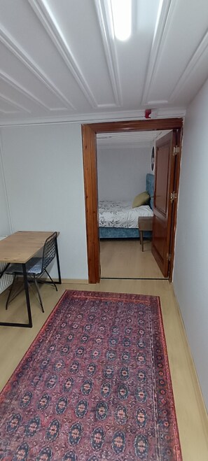 Une pièce avec une table en bois, un tapis à motifs, un lit avec une tête de lit bleue et une porte donnant sur une autre pièce.